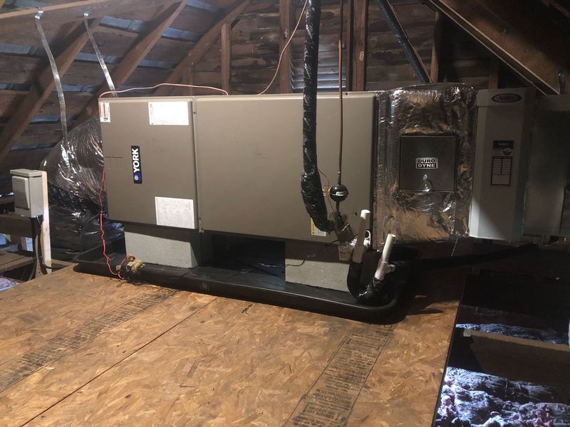 HVAC unit in an attic. Large, rectangular metal unit with ductwork and insulation, set on a platform.