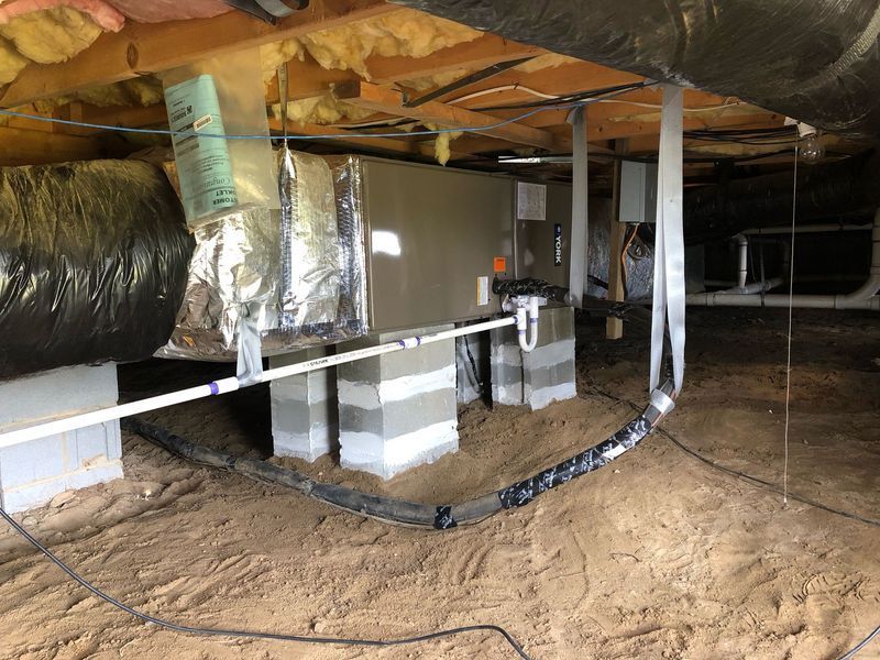 HVAC unit in a crawl space, supported by concrete blocks, with ductwork and piping visible.