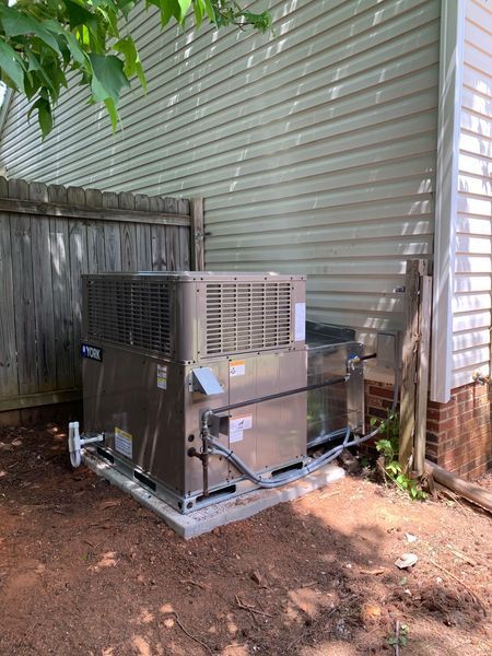 HVAC unit on a concrete pad between a fence and a building. Brown siding, red dirt.