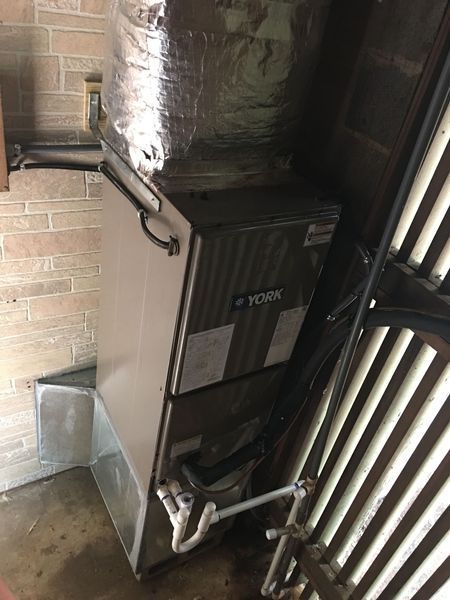 York furnace, silver, in a brick-walled utility room, with attached ductwork and pipes.