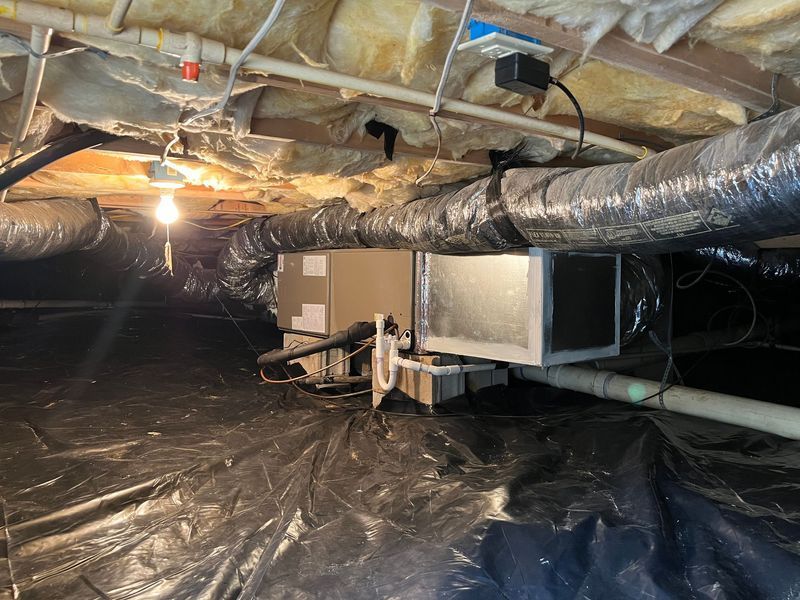 Crawlspace with HVAC unit, ductwork, and insulation. Black plastic ground cover, dim lighting, pipes, and electrical components.