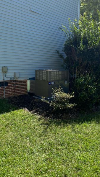 An air conditioning unit next to a light blue house and a green bush, on a patch of grass.
