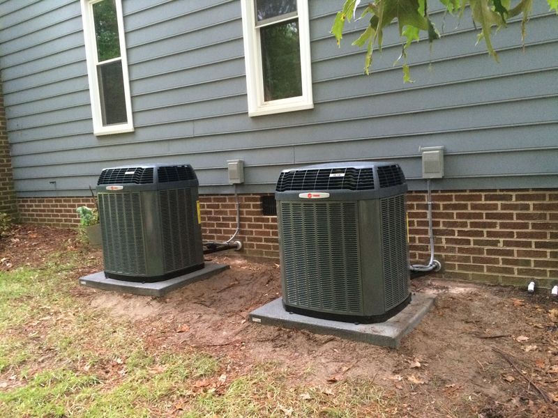 Two gray air conditioning units outside a gray-sided house with brick base and two windows.