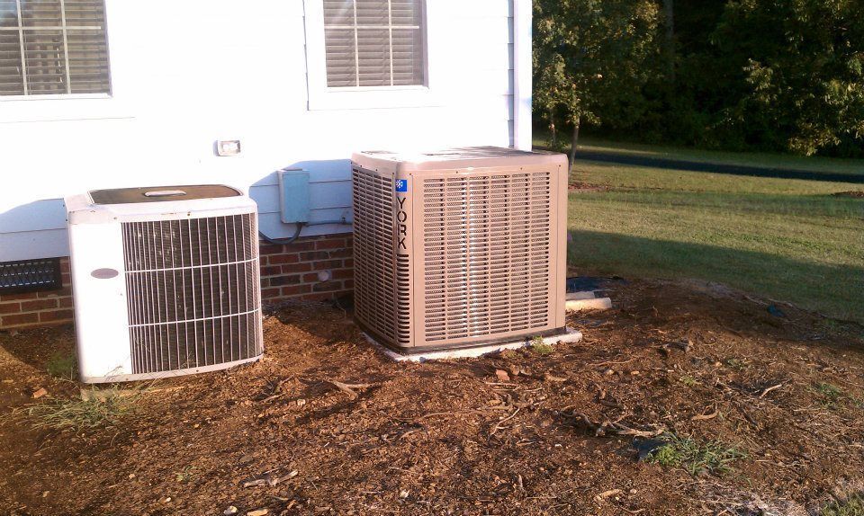 Two air conditioning units sit outside a white building with windows, on a patch of brown soil.