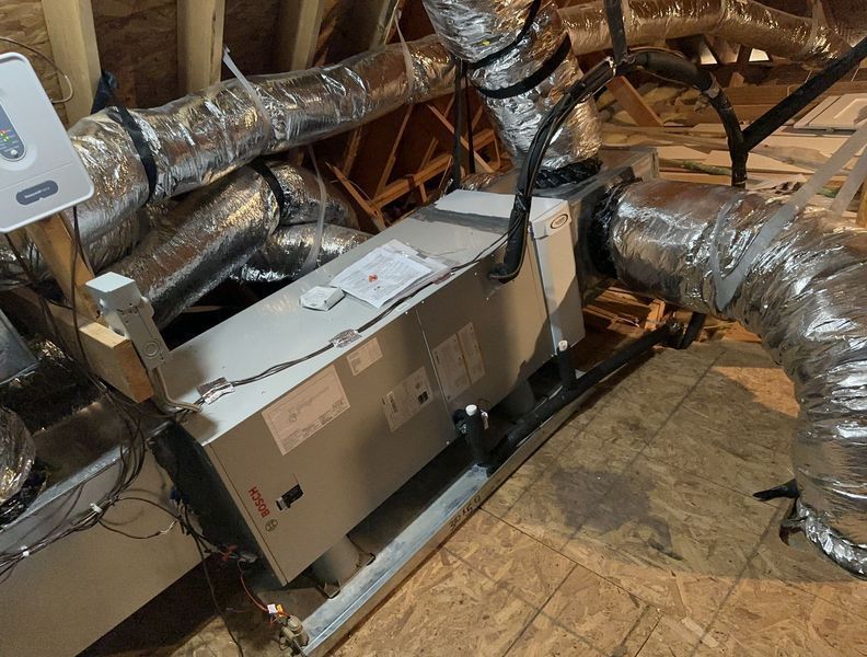 HVAC system in an attic. Ductwork connects to a large, rectangular unit. Wires and a control panel are also visible.