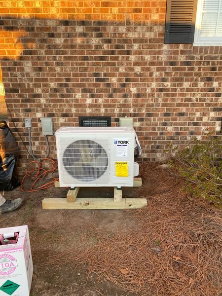 York air conditioning unit on a wooden base against a brick wall.  Electrical connections nearby, with a person present.