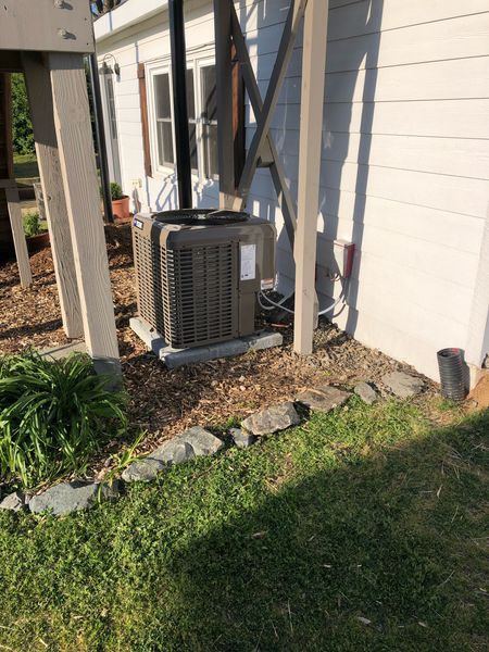 An outdoor air conditioning unit sits near a white house with a wooden support structure.