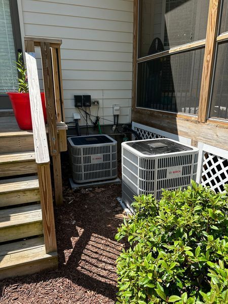 Two air conditioning units next to a wooden deck and a house with a window.