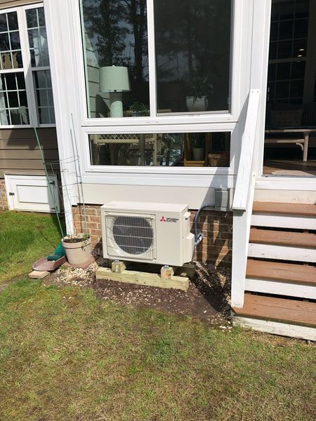 Mitsubishi heat pump unit installed outside, below a window. On a wooden base, near grass and stairs.