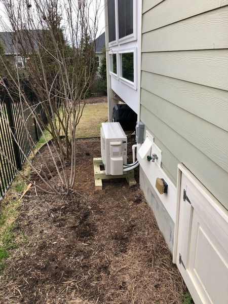 HVAC unit on wooden supports next to a light-colored house. Dark mulch surrounds the unit and a bare tree.
