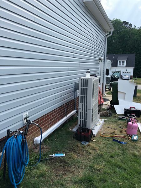 HVAC unit installed near a house exterior with light blue siding. Yard tools and hoses are nearby.