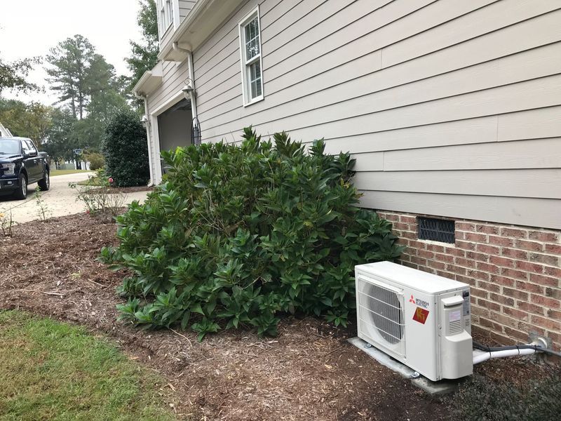 Exterior view of a house with beige siding, shrubbery, and a white air conditioning unit on a brick foundation.