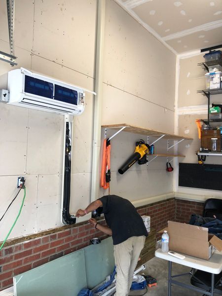 Person installing HVAC unit in a garage. Air conditioner mounted on wall, brick base, white walls.