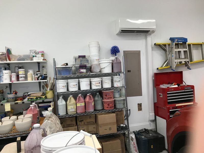 A workshop with shelves holding paint buckets and tools, a ladder, and an air conditioner.