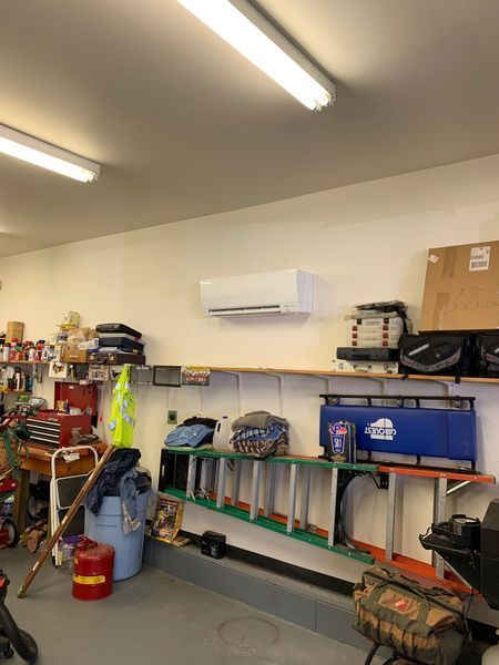 White air conditioning unit mounted on a white wall in a garage. Shelves hold tools, containers, and a ladder.