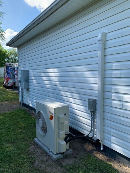 White heat pump unit on a concrete pad next to a house with white siding and electrical boxes.