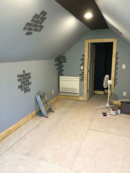 Room with gray walls, unfinished floor, air vent, door, fan, ladder, and brick stencil designs.
