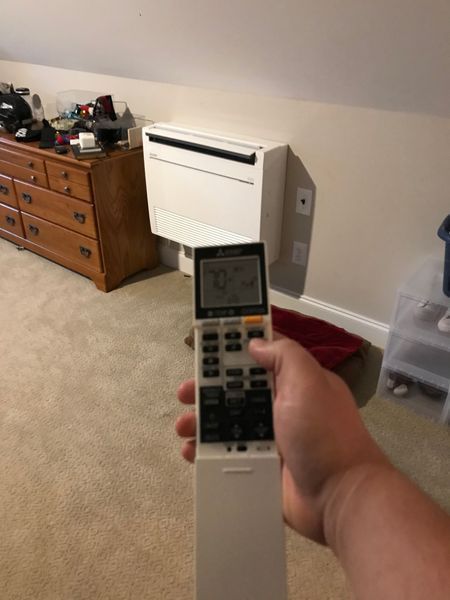Hand holding a remote control pointed at a wall-mounted white air conditioning unit.