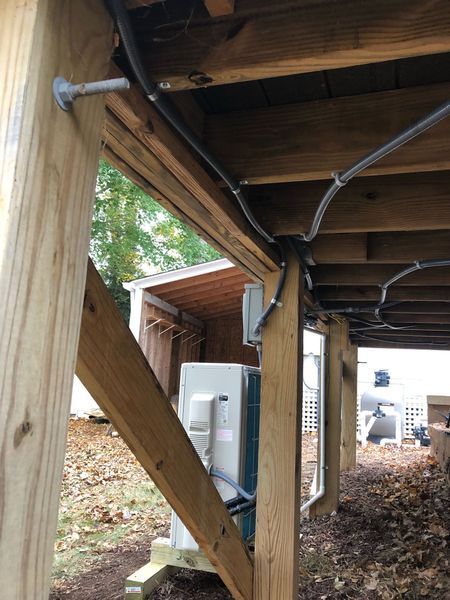 Underneath a wooden deck, electrical wiring runs along beams. An HVAC unit is visible, with a small shed in the background.