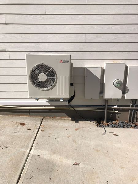 Mitsubishi heat pump unit mounted on a white house exterior with electrical boxes and wiring.