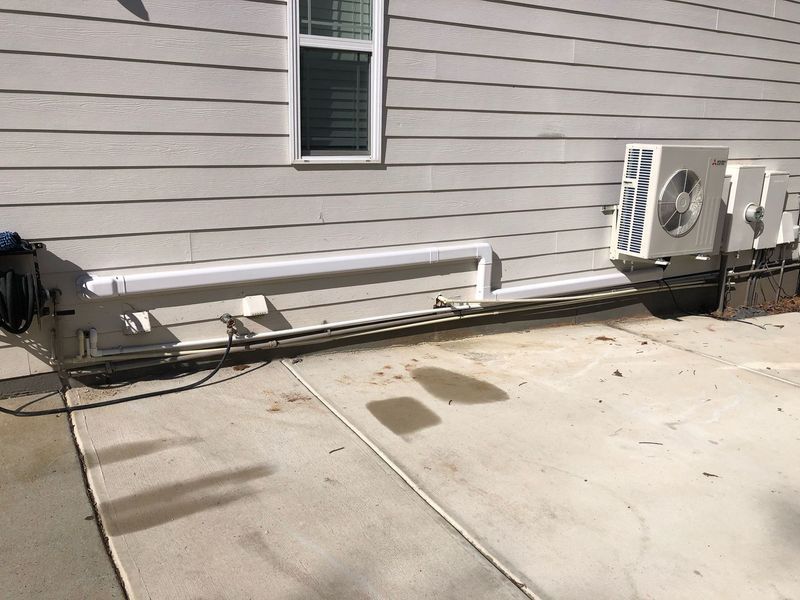 Air conditioning unit mounted on a house exterior. White siding, concrete patio, and pipe conduit visible.