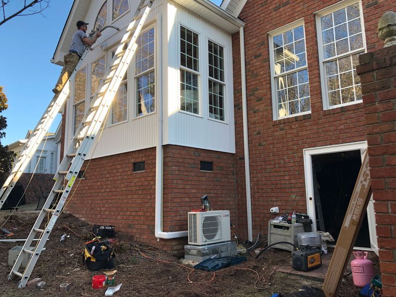 Man on ladder, installing AC unit on a brick building with white trim and windows. Tools and equipment on the ground.