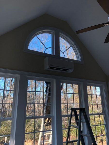 An air conditioning unit is installed below a semi-circular window. White-framed windows span the lower wall, with a ladder in view.