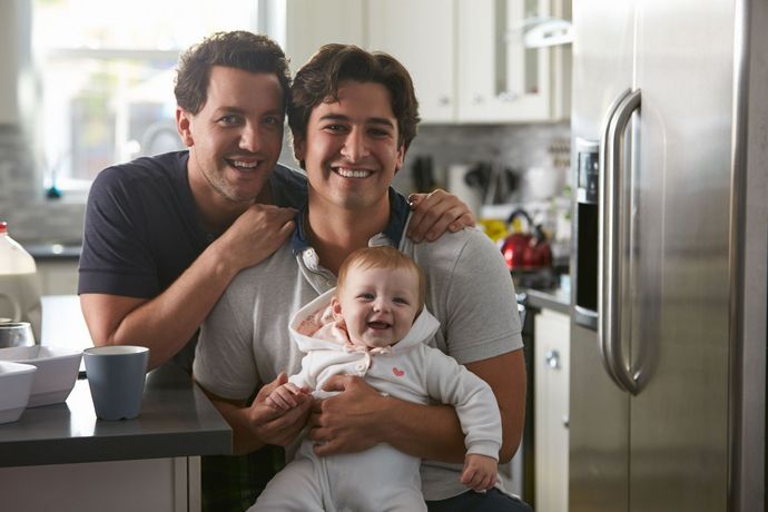 Two men are holding a baby in a kitchen.