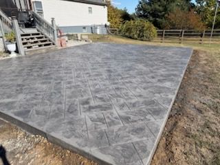 Gray stamped concrete patio next to a house with wooden stairs, surrounded by grass.