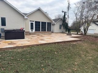 Backyard patio with a hot tub, fire pit, and a house in the background. Green grass and overcast sky.