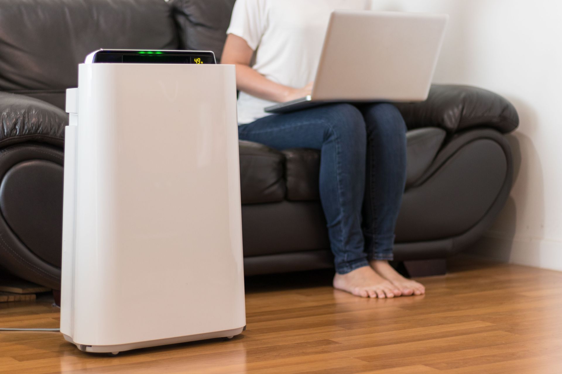 A tall, white air purifier stands on a wood floor next to a person sitting on a leather sofa using a laptop.