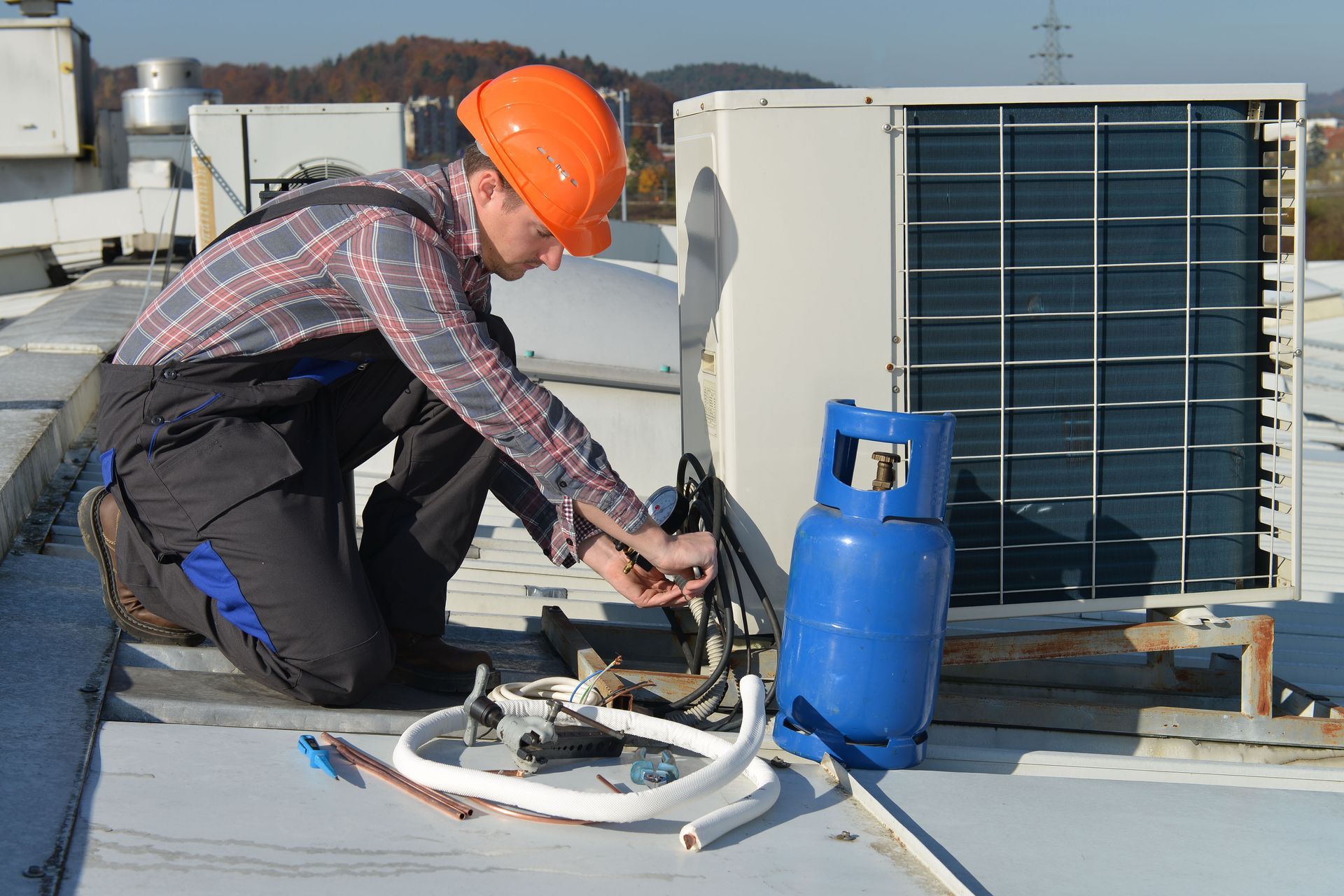 A technician in a hard hat kneeling on a rooftop while servicing an air conditioning unit next to a blue gas cylinder.