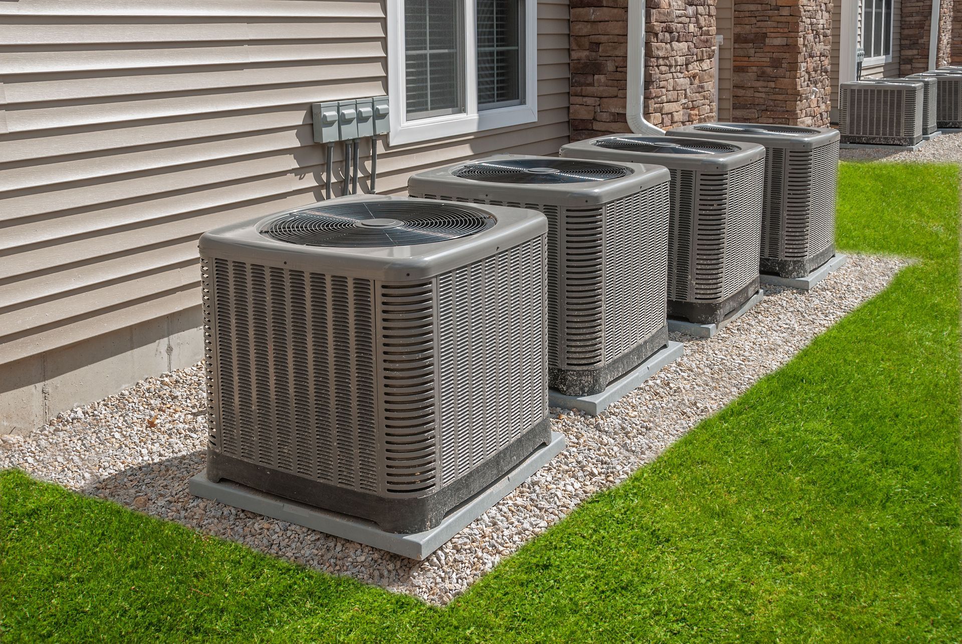 Four air conditioning units sit on a gravel path next to a beige-sided building and a patch of green grass.
