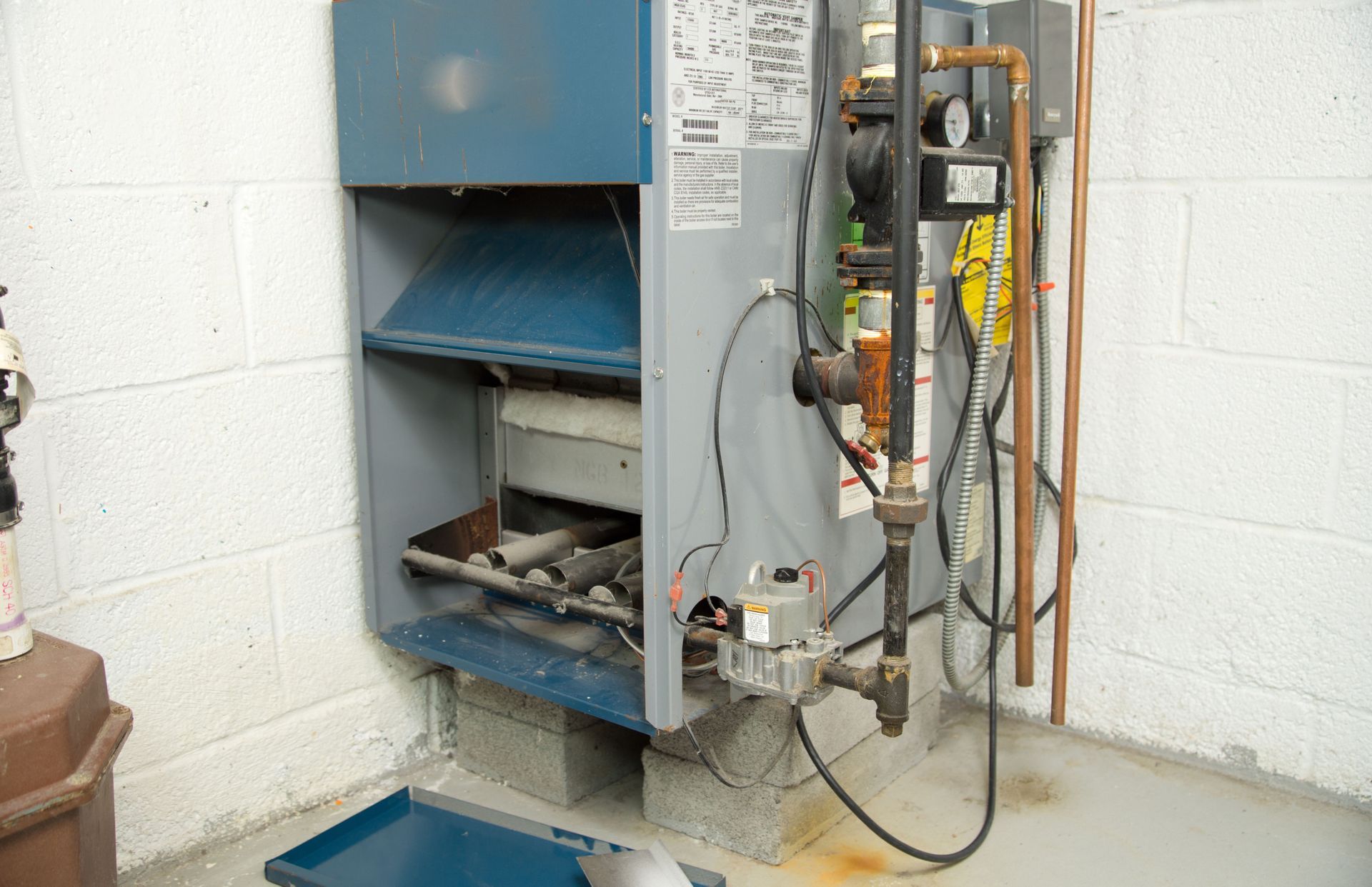 A grey furnace with a blue panel sits on cinder blocks against a cinder block wall in a utility room.