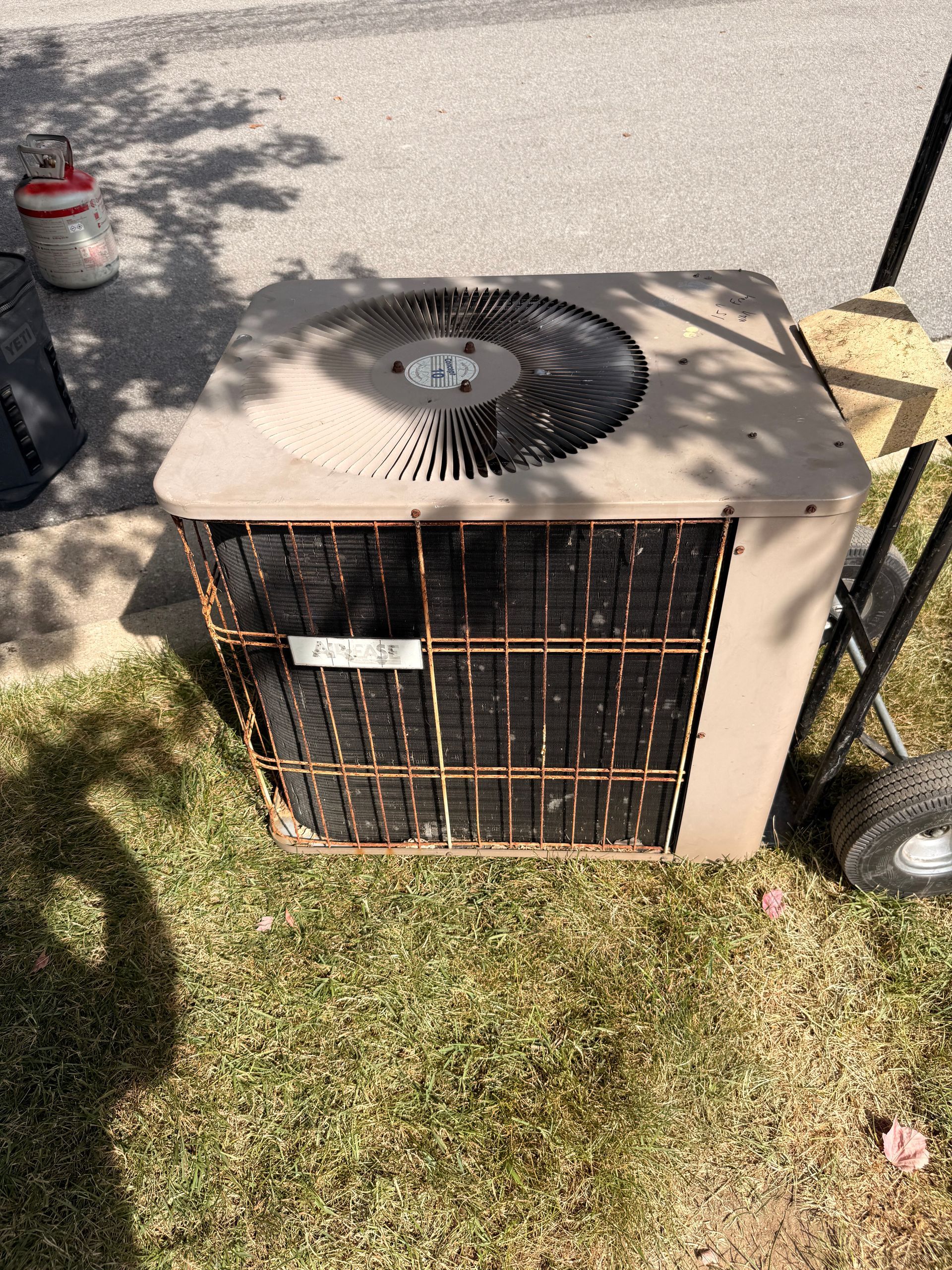 A tan, square outdoor air conditioner unit sits on a grass lawn next to a metal hand truck.