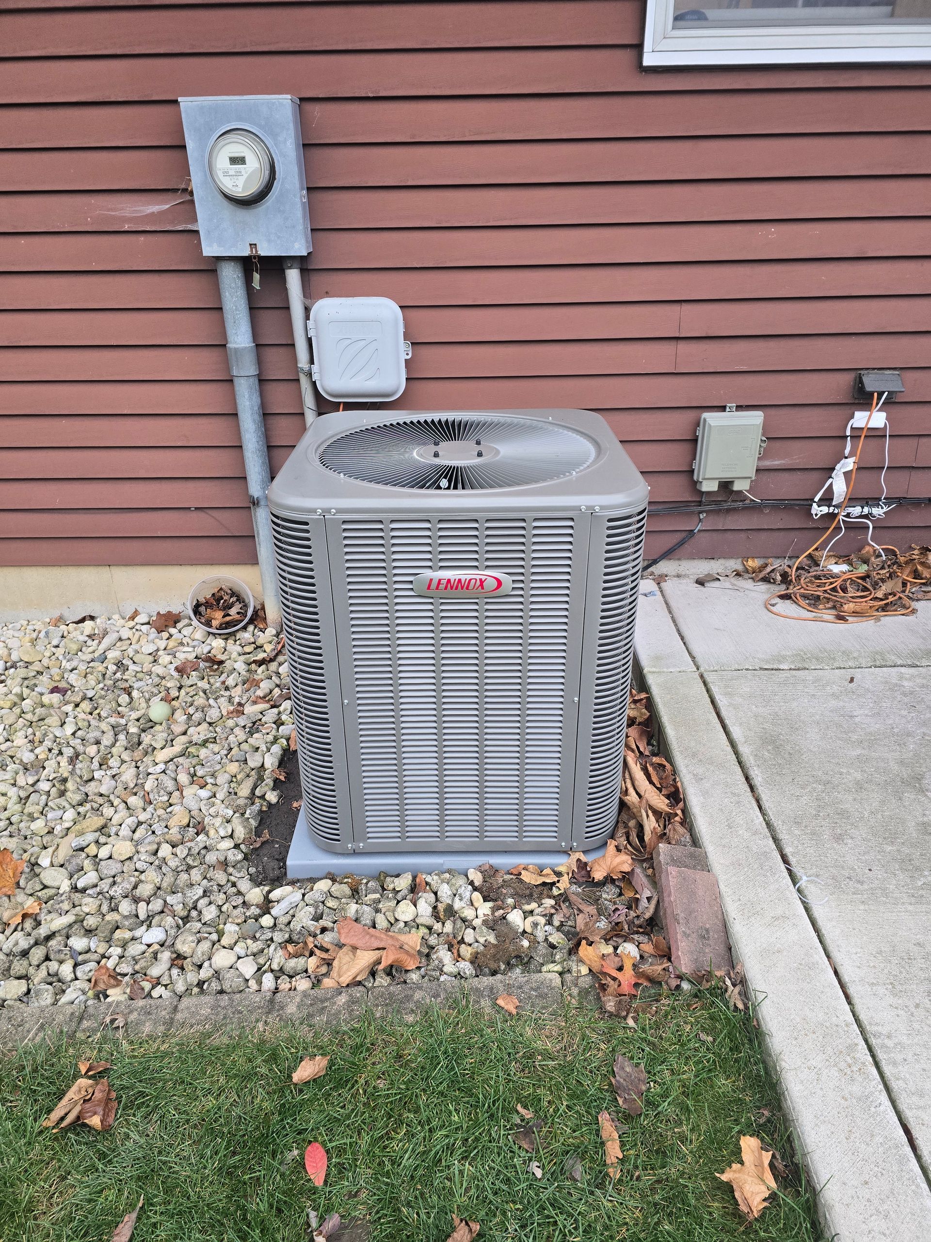 A grey Lennox central air conditioning unit sits on a concrete slab against the side of a red-sided house.