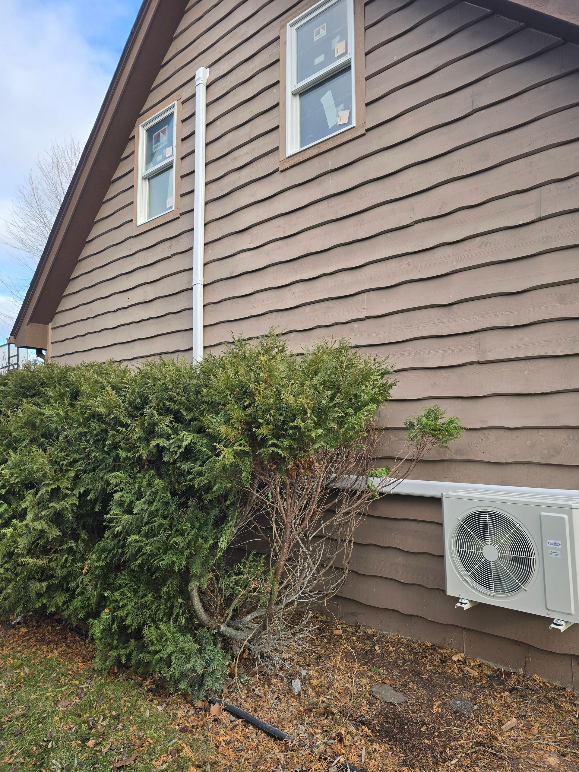 Exterior view of a brown-sided house with two windows, a white vertical pipe, and an outdoor HVAC unit near a bush.