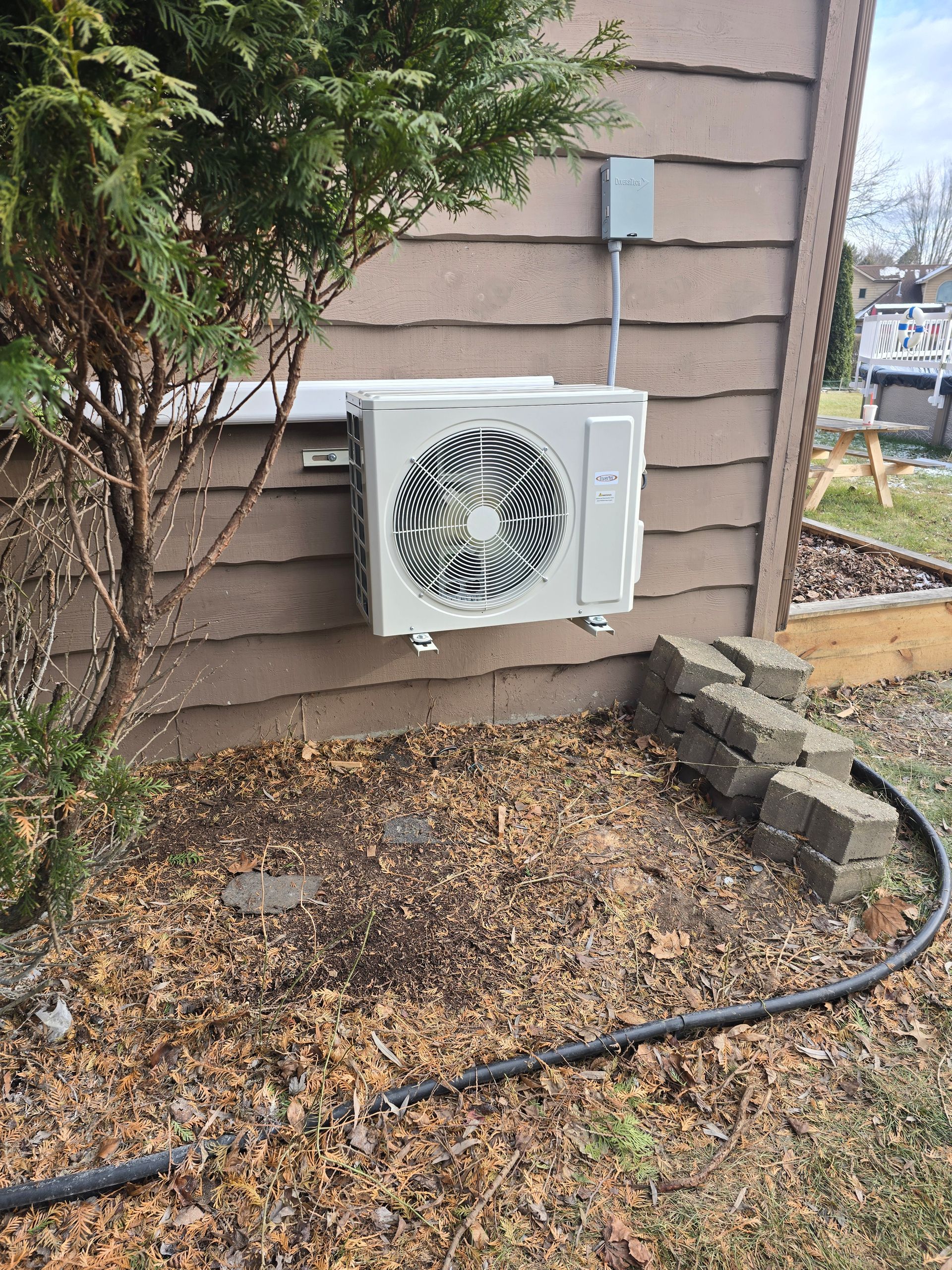 A white mini-split air conditioning unit mounted on the exterior siding of a house, next to a bush and loose garden bricks.
