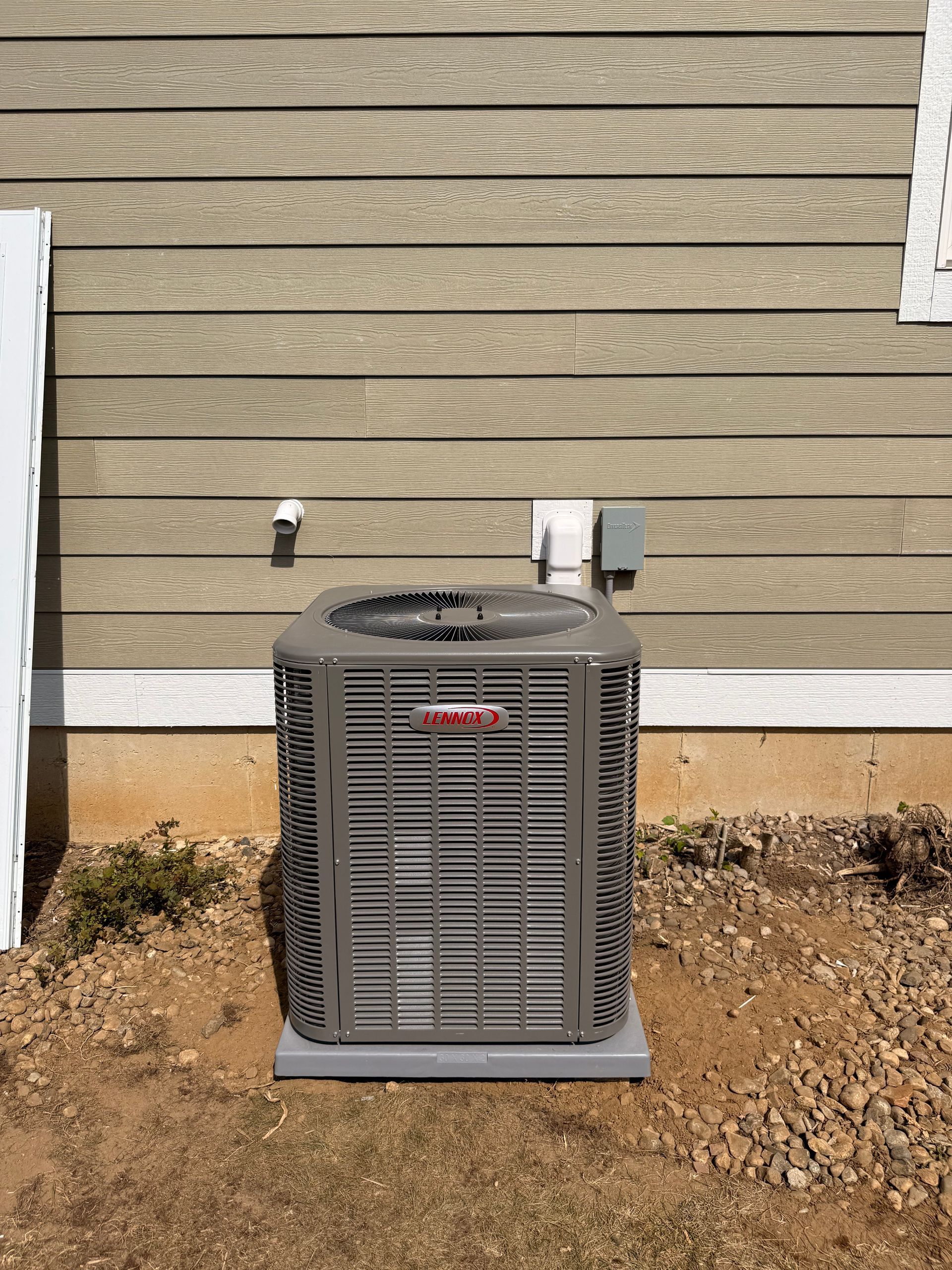 A gray Lennox air conditioning unit sits on a gravel patch against a tan horizontal siding house wall.