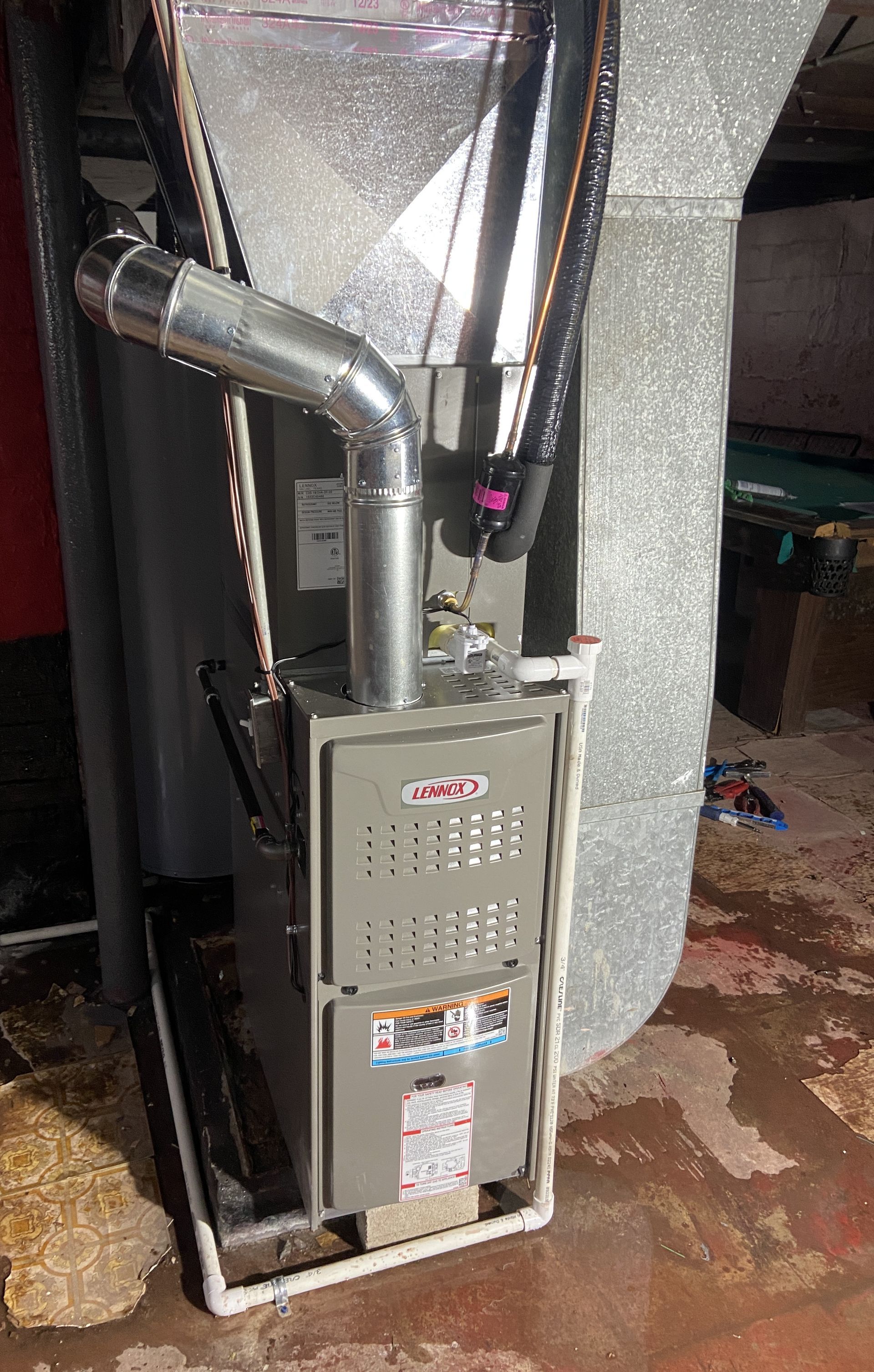 A gray HVAC furnace unit stands on a white frame in a basement, connected to silver ductwork and white PVC piping.