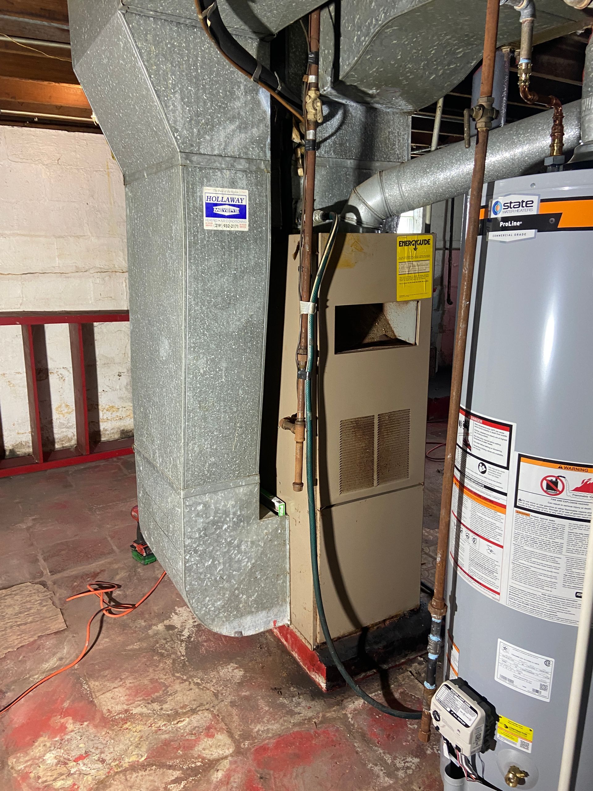 A tan furnace stands next to a gray water heater in a basement with metal ductwork, red flooring, and a stone wall.