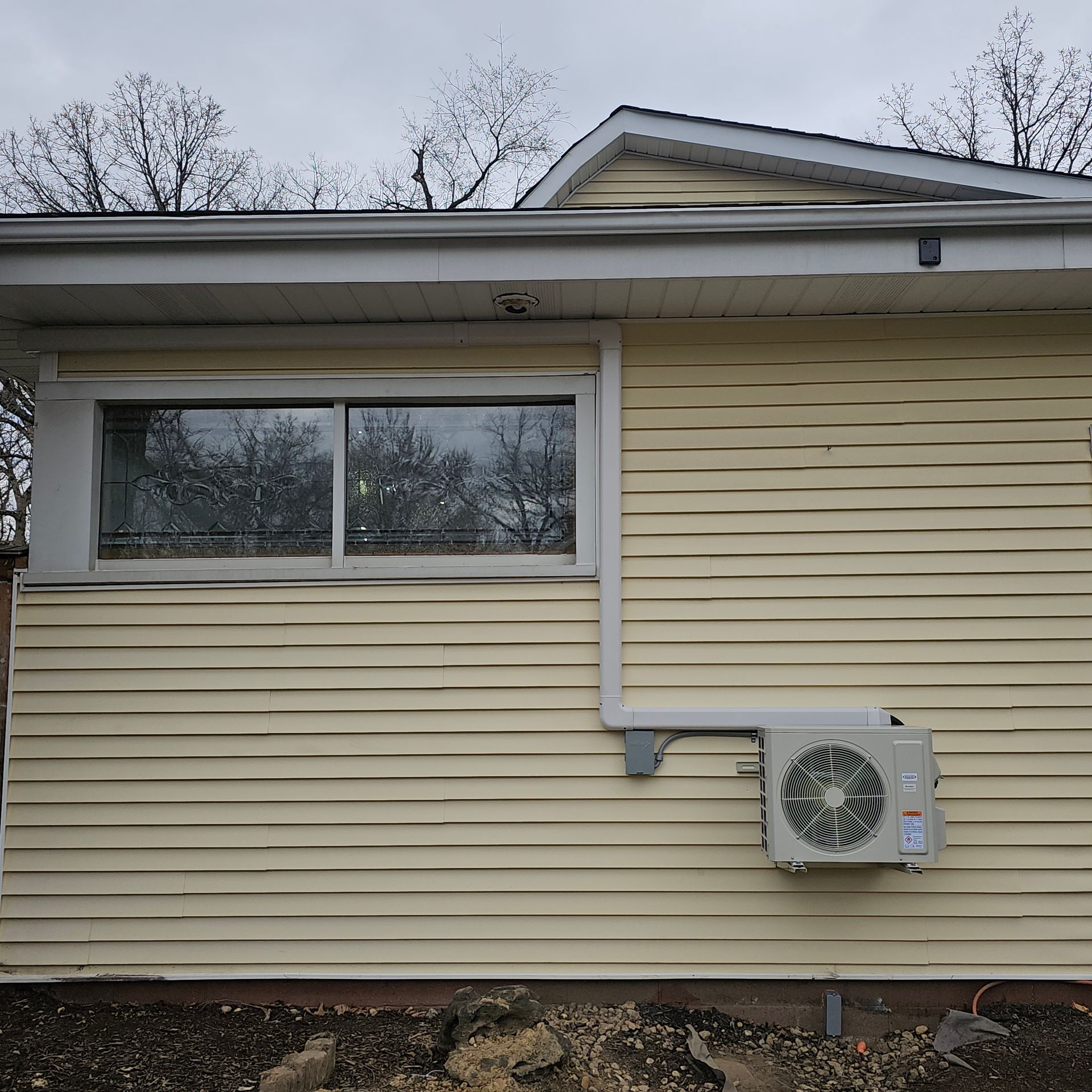 Light yellow horizontal siding on a house exterior featuring a sliding window and an outdoor HVAC unit mounted on the wall.