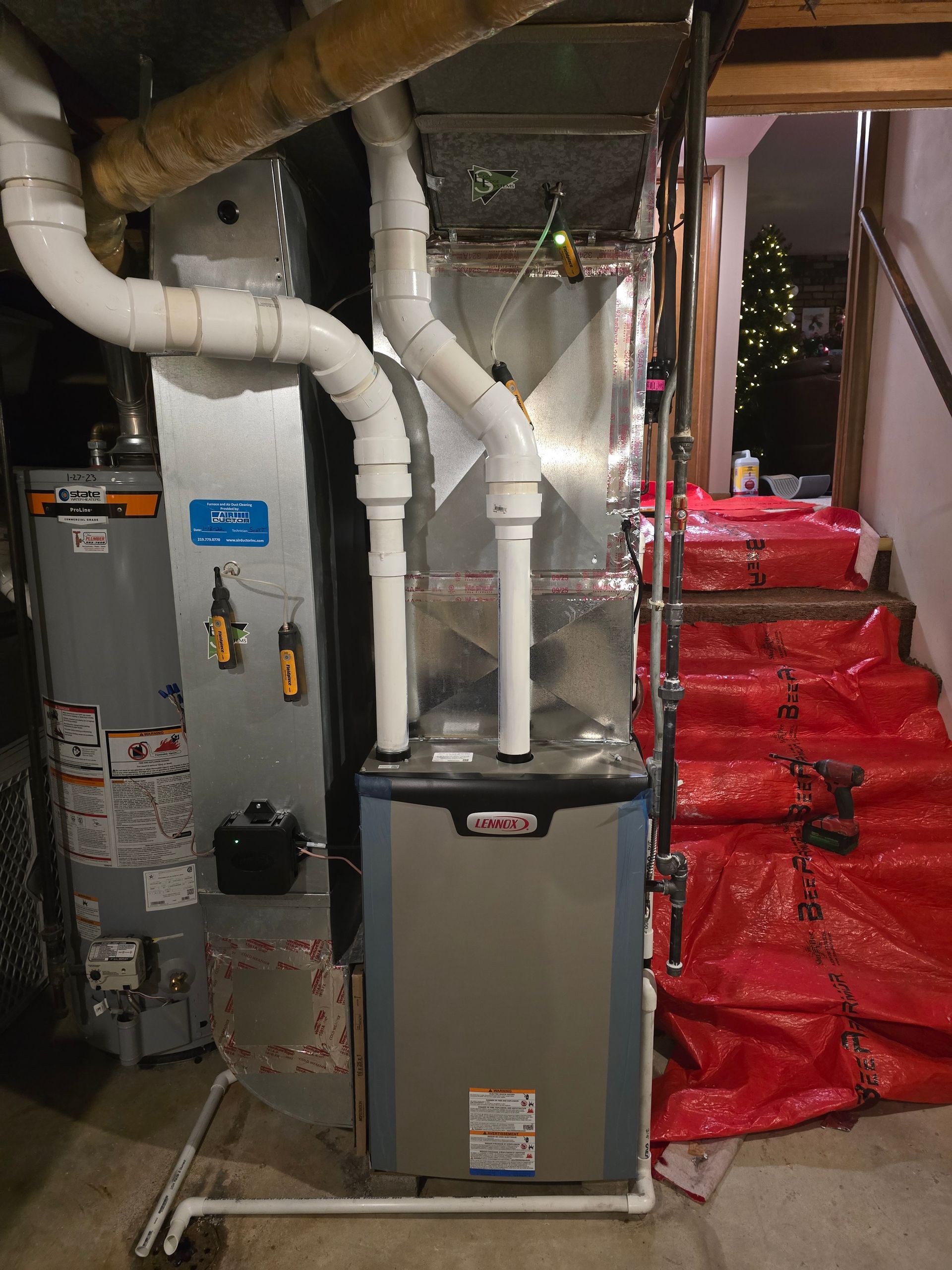 A high-efficiency gas furnace and water heater installation in a basement, with red protective plastic on nearby stairs.