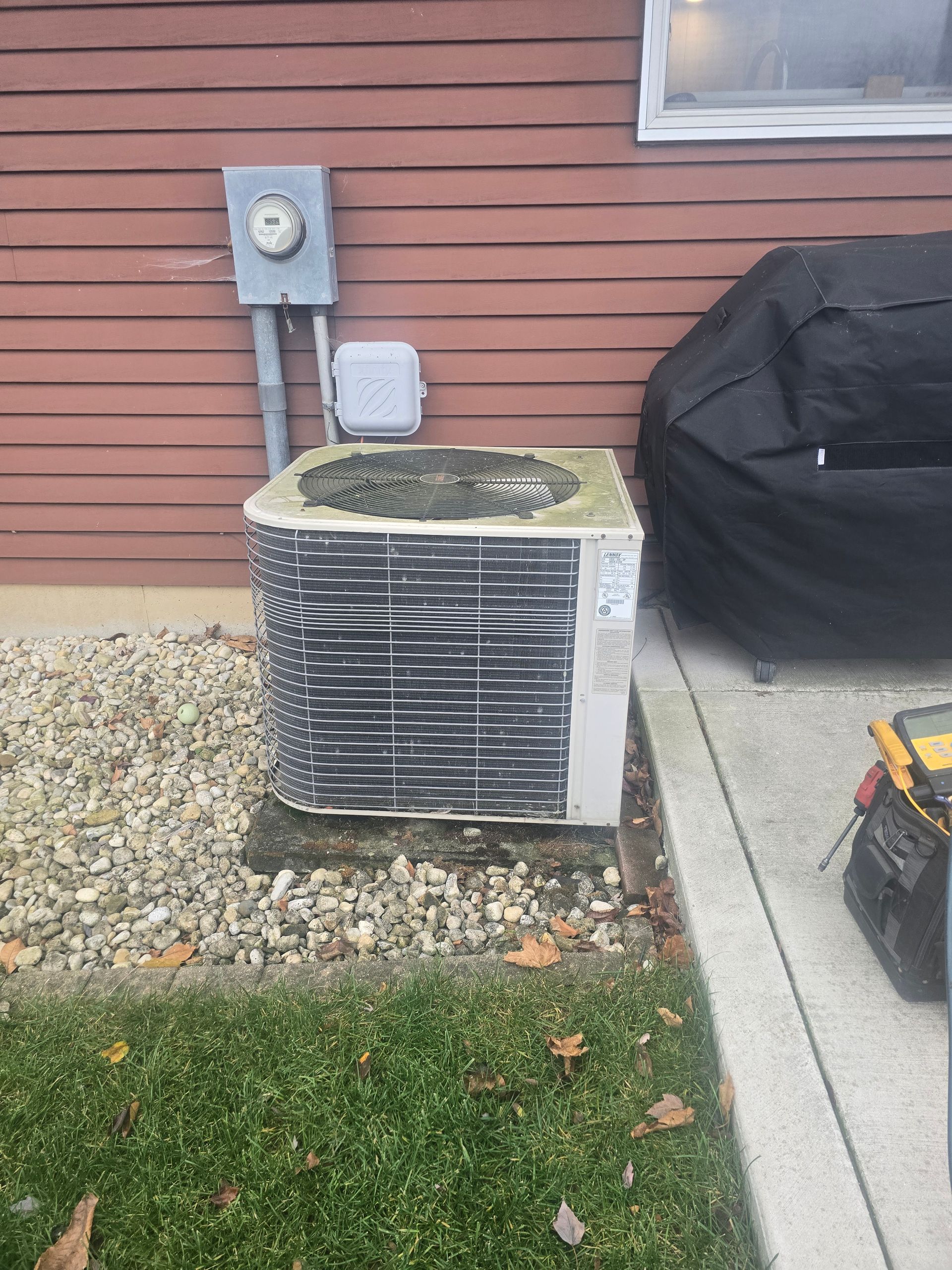 An outdoor air conditioning unit sits on a gravel patch beside a brown house, with a grill cover and tools nearby.