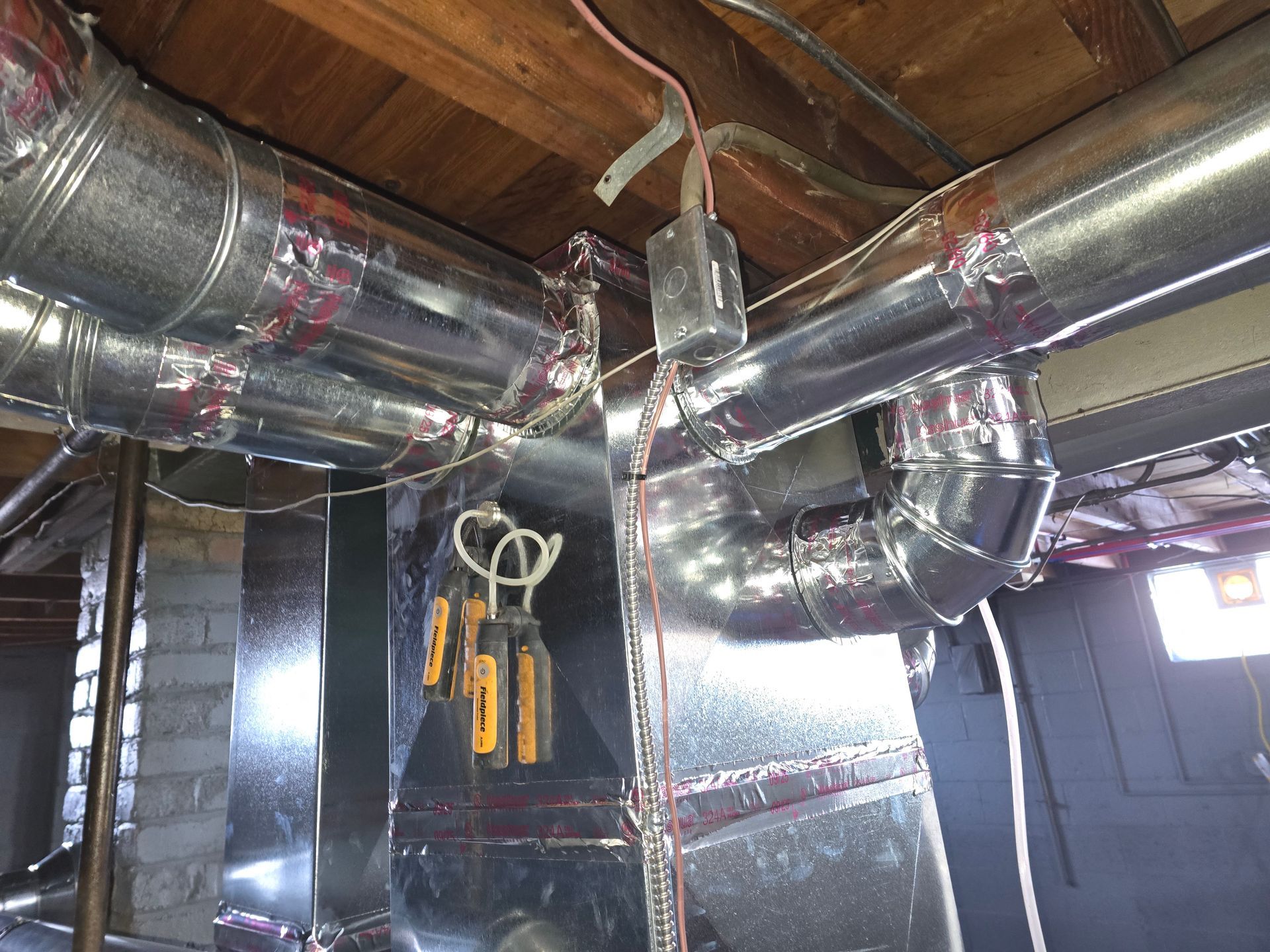 Metallic HVAC ductwork and a gray junction box are installed on wooden ceiling joists in a basement setting.