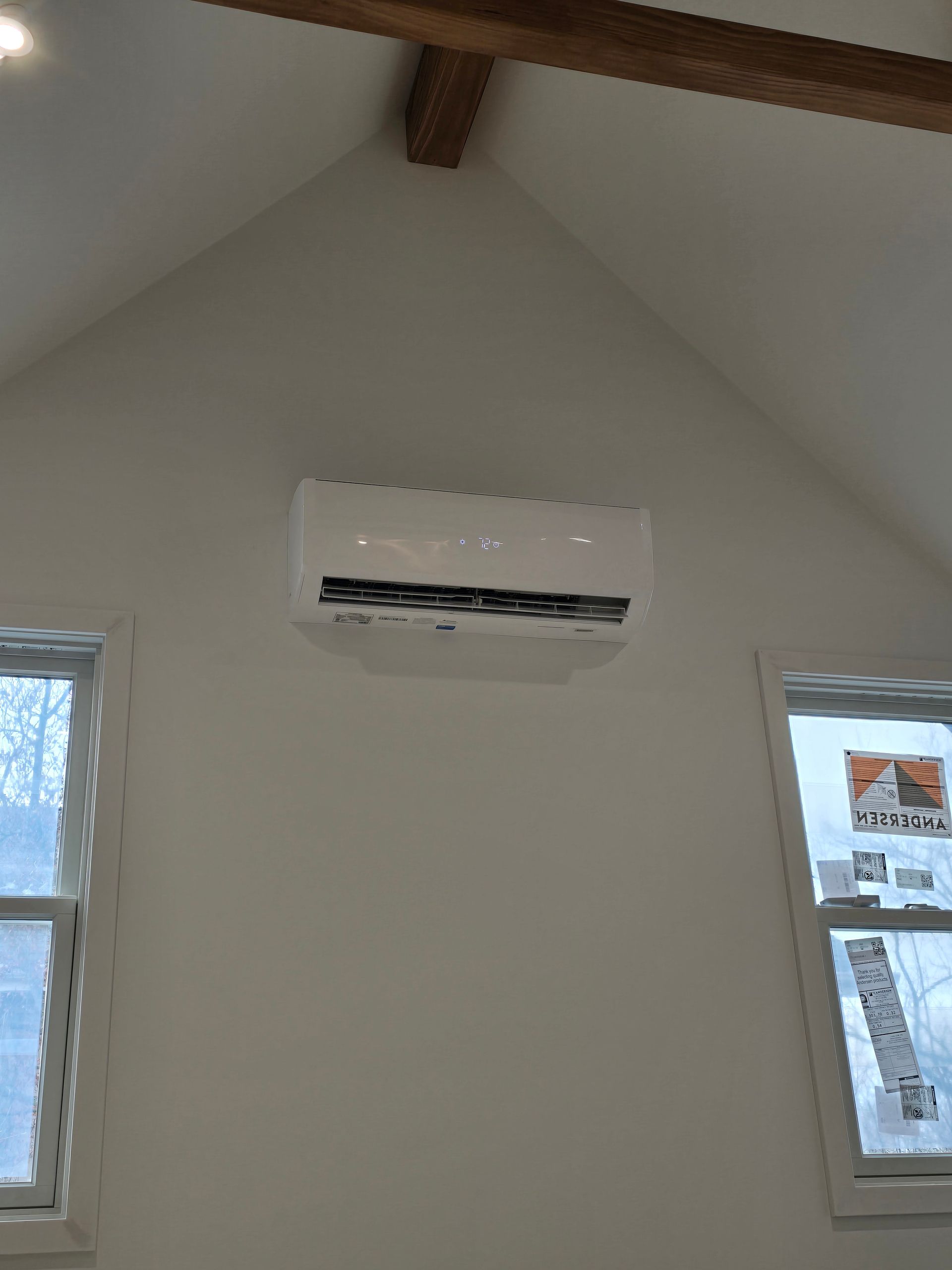 A white ductless mini-split air conditioner mounted on a beige wall between two windows under a vaulted ceiling.