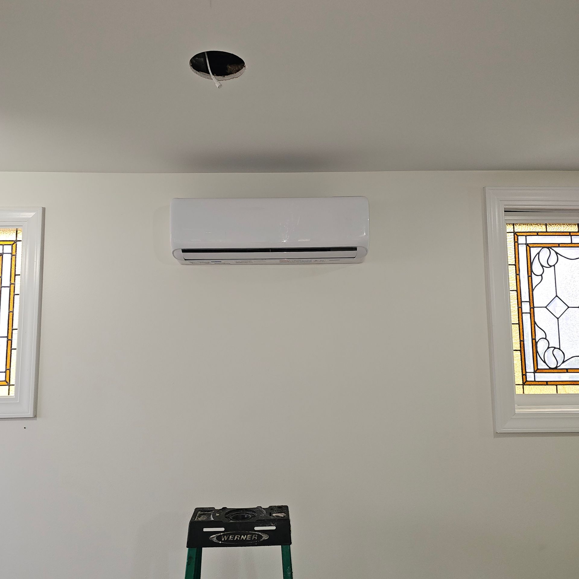 A wall-mounted white air conditioning unit installed between two decorative stained-glass windows in a room.