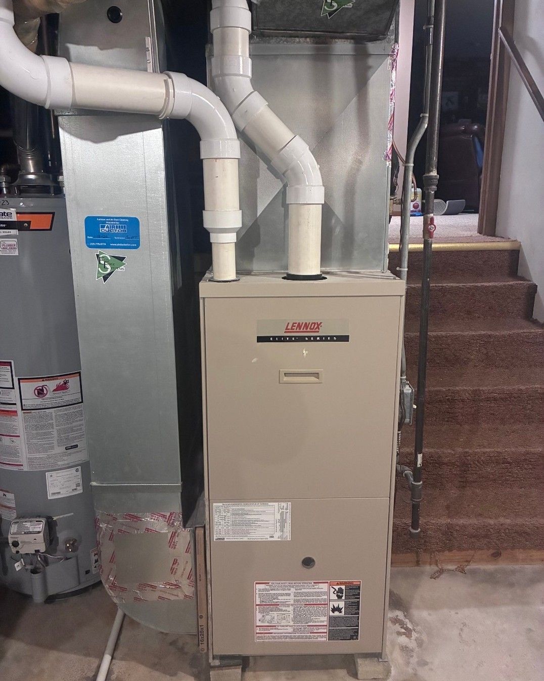 A beige gas furnace and water heater with white PVC venting pipes in a basement next to carpeted stairs.