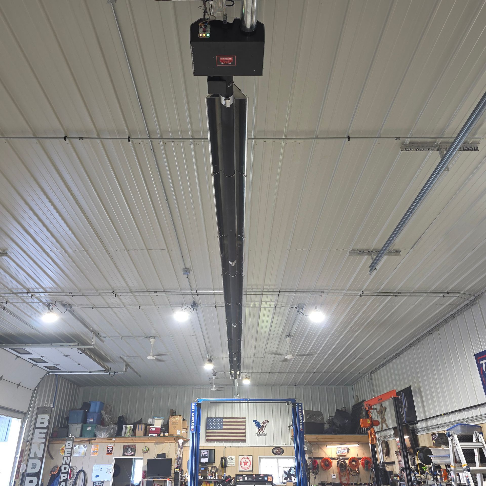 A black tube heater mounted to the ceiling of a cluttered garage workshop.