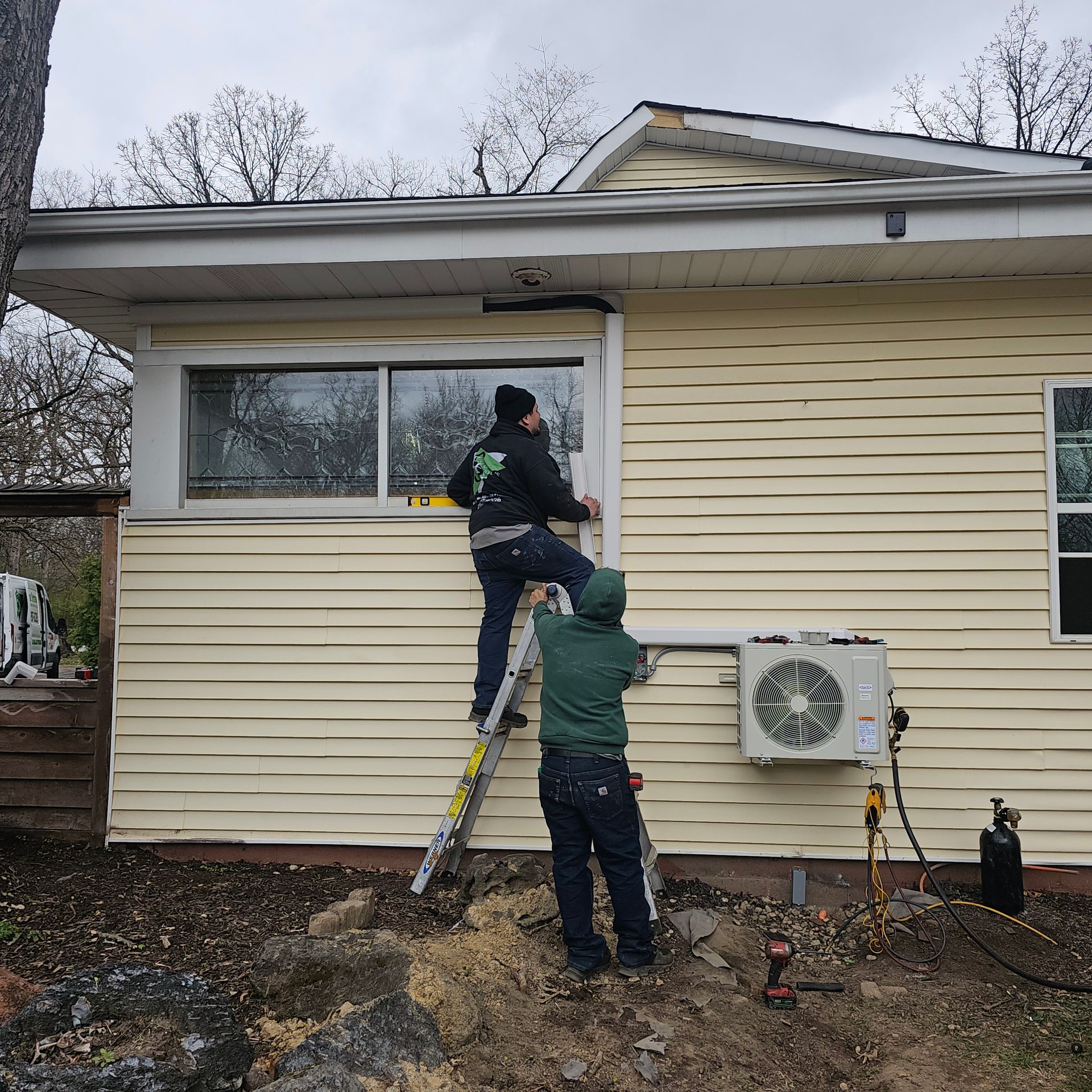 Two people in casual work clothing use a ladder to install or adjust a window on the side of a light yellow, sided house.